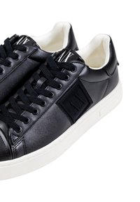 Zapatillas negras de cuero sintético con diseño de cordones, suela de goma blanca, acentos texturizados y logo en el costado. Estilo minimalista.