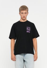 Young man wearing a black oversized t-shirt with a purple mountain graphic on the chest, paired with light blue jeans, standing against white background.