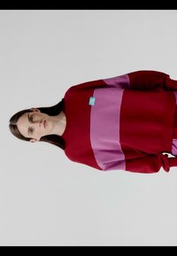 Ung person med langt mørkt hår iført en rød og pink stribet sweatshirt med et lille blåt mærke på brystet, stående mod en ensfarvet baggrund.