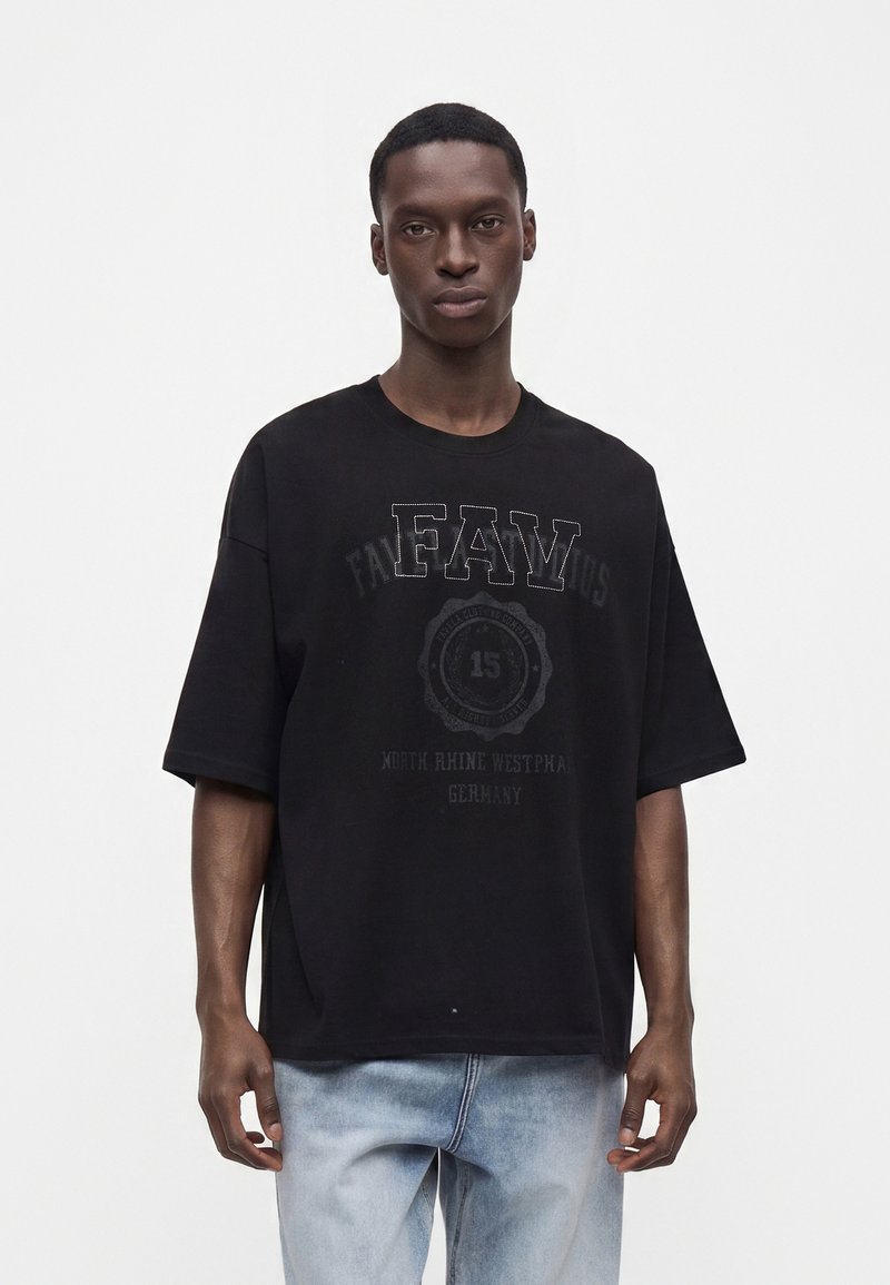 Νέος άνδρας φοράει μαύρο oversized t-shirt με ξεθωριασμένο κείμενο και ανοιχτόμπλε τζιν, στέκεται μπροστά σε ένα απλό λευκό φόντο.