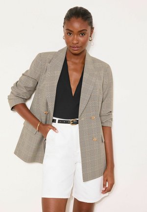 Nő, bézs kockás blézert visel fekete V-nyakú felsővel, fehér rövidnadrággal és arany karika fülbevalókkal, egyik kezét zsebre dugva áll.