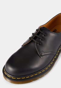 Chaussure en cuir noir avec bout rond, lacets sur le devant et piqûres jaunes le long de la semelle. Texture lisse et doublure intérieure en brun contrastant.