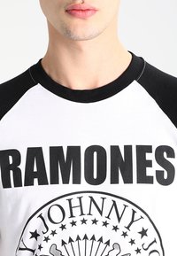 Camiseta blanca con mangas raglán negras, que presenta un texto negro en negrita "RAMONES" y un diseño gráfico circular con estrellas y texto debajo.