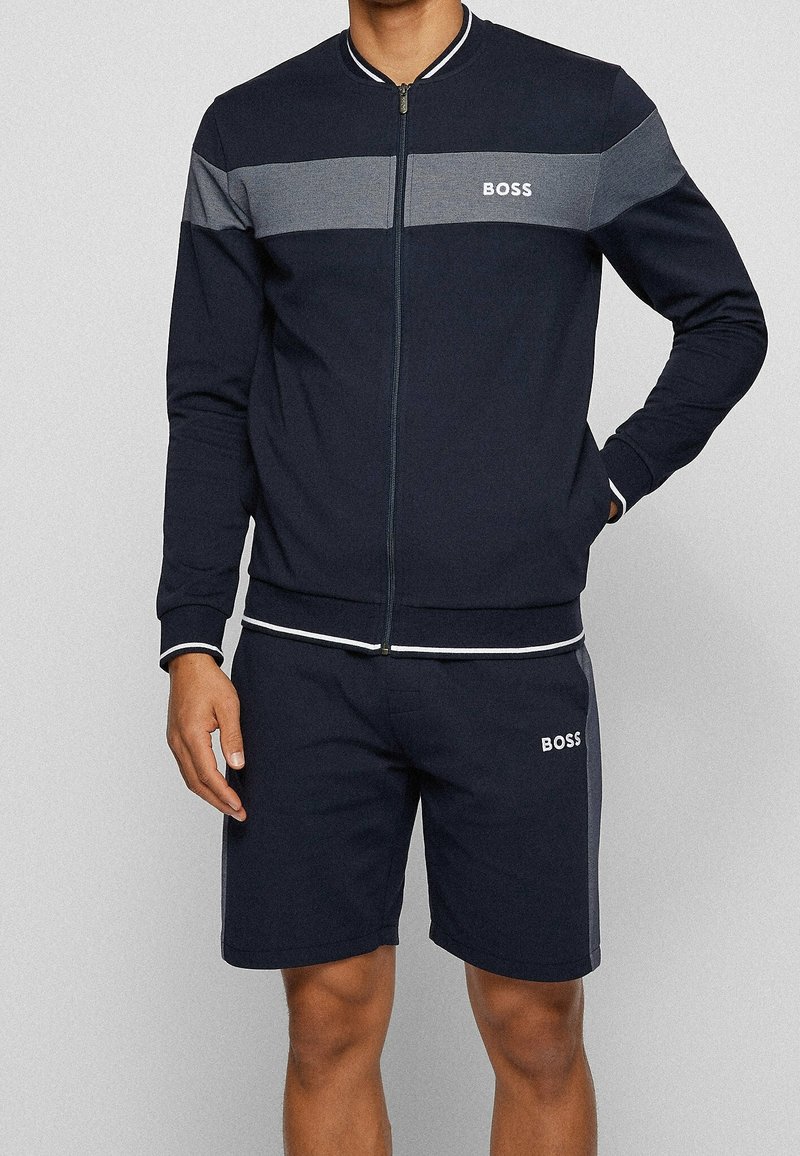 Marinblå atletisk träningsdräkt med en grå randig panel, som har en zip-up jacka och matchande shorts. Logotyp på bröstet och låret. Blandning av bomullsmaterial.