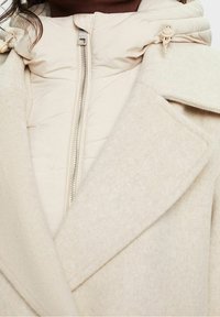 Abrigo de mezcla de lana beige con una capa interior tipo plumón. Cuenta con un cierre frontal, grandes solapas y cordones ajustables en la capucha.