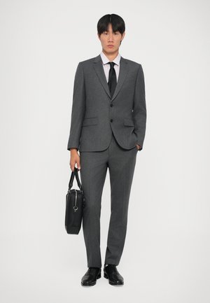 Completo grigio su misura con due bottoni, tasche a pattina e una cravatta nera. È visibile una camicia rosa chiaro. Le scarpe nere e una borsa nera completano il look.