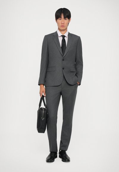 Costume gris ajusté avec deux boutons, rabats de poche et une cravate noire. Une chemise rose clair est visible. Des chaussures noires et un sac noir complètent le look.