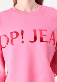 Pink sweatshirt with a crew neck featuring a glossy red print of the text "POP! JEAN." The fabric appears soft with a smooth texture.