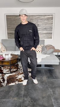 Juodas pūkuotas megztinis su apvaliu kaklu, derinamas su juodomis kelnėmis, turinčiomis kelio plyšius. Baltos sportinės bateliai. Neutraulios spalvų tonų baldai fone.