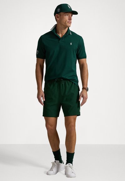 Tricou polo verde cu un guler cu dungi contrastante, pantaloni șort verzi asortați și adidași albi. Prezintă un logo pe tricou și pe șapcă.