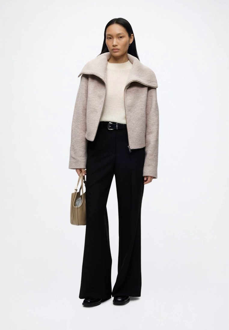 Femme portant une veste beige zippée avec un large col, un pull crème, un pantalon noir à jambes larges, des chaussures noires et tenant un sac à main marron clair.
