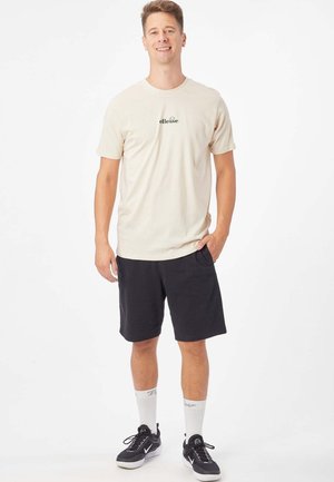 Camiseta de algodón beige claro con logo verde, combinada con pantalones cortos negros. Zapatillas negras y calcetines blancos con detalles grises completan el conjunto.