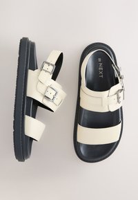 Sandales à semelle noire avec larges brides crème et boucles argentées, vues de dessus sur un fond clair.