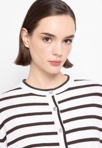 Cardigan bianco con strisce orizzontali marroni. Presenta un collo rotondo, chiusura con bottoni e una texture in maglia liscia.