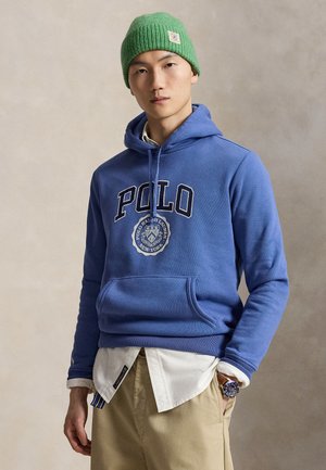 Młody mężczyzna w zielonej czapce typu beanie, niebieskiej bluzie Polo z kapturem i logo, białej koszuli pod spodem, beżowych spodniach oraz zegarku na rękę, stojący na jednolitym tle.