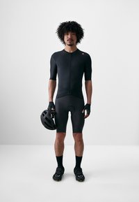 Schwarzer Radanzug mit kurzen Ärmeln, Reißverschluss vorne und gepolsterten Shorts. Matte Textur, elegantes Design. Fahrer trägt schwarze Handschuhe und hält einen Helm.