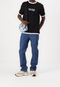 Zwart T-shirt met witte kraag en grafische tekst, blauwe spijkerbroek, beige sneakers en een lichtblauwe schoudertas met zwarte accenten.