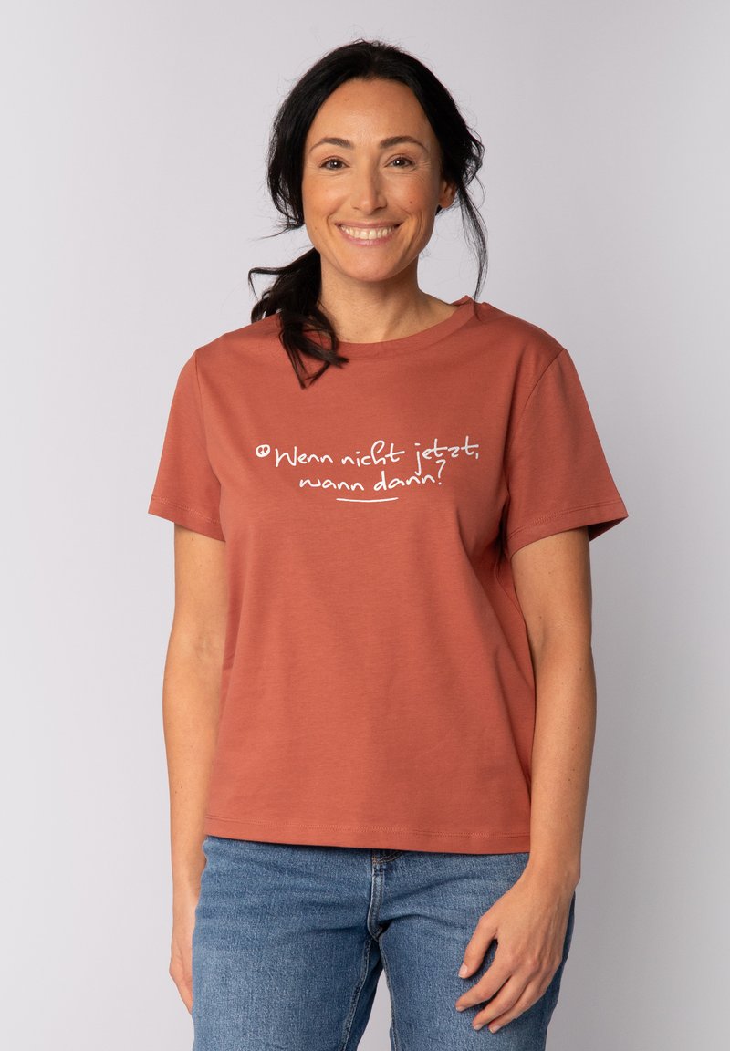 Eisenbraunes T-Shirt aus weichem Stoff mit kurzen Ärmeln. Verfügt über einen weißen Drucksatz auf der Brust. Kombiniert mit blauen Jeans.