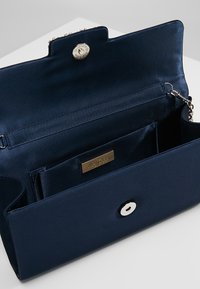 Námořnická saténová kabelka na ruku se zacvakávacím uzávěrem, stříbrným kováním a vnitřní kapsou. Má texturovaný povrch a detaily s logem.