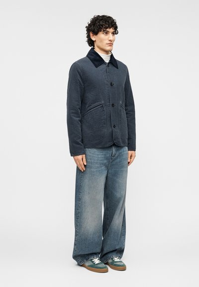 Ναυτικό μπουφάν με καπιτονέ υφή, κλείσιμο με κουμπιά και δύο εμπρόσθιες τσέπες. Συνδυασμένο με φαρδιά μπλε τζιν και πράσινα αθλητικά παπούτσια.