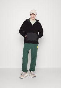 Pullover in pile nero con zip a metà e grande tasca nera, abbinato a pantaloni della tuta verdi con logo e sneakers chiare.