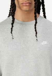 Lys grå Nike-genser med rund hals, korte ermer og en liten hvit logo på brystet. Mykt, teksturert stoff.