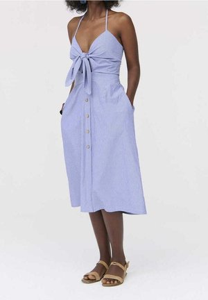 Vrouw die een midi-jurk draagt met blauwe en witte strepen, knopen aan de voorkant, een gestrikte bustier, dunne bandjes en beige sandalen, staand met handen in de zakken.