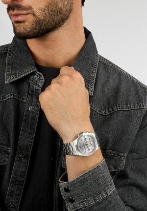 Silberner Chronograph mit weißem Zifferblatt, Edelstahl-Armband und Akzenten. Am Handgelenk getragen, teilweise von einer Jeansjacke bedeckt.