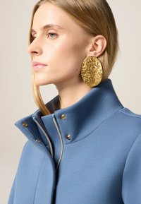 Giacca blu con colletto alto, dotata di dettagli in metallo dorato e zip. Orecchini grandi e testurizzati in oro aggiungono contrasto. Tessuto morbido con una finitura pulita.