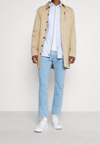 Beżowy długi płaszcz, jasnoniebieska koszula na guziki, jasnoniebieskie jeansy oraz białe trampki z zielonymi akcentami. Casualowa, warstwowa stylizacja.