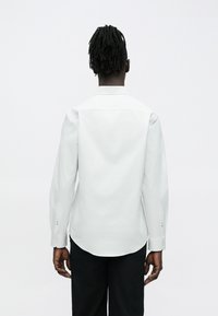 Homme aux courtes dreadlocks portant une chemise blanche impeccable à manches longues et un pantalon noir, debout de dos sur un fond clair uni.