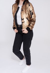 Giacca bomber in paillettes dorate con dettagli neri, chiusura frontale con zip e tasche laterali. Indossata con pantaloni in denim neri e sneakers bianche. Design minimalista.
