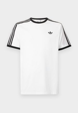 Balts kokvilnas t-kreklis ar melnu ribotu kaklu un piedurknēm, kuram uz katra pleca ir trīs melnas svītras un neliels logo krūtīs.