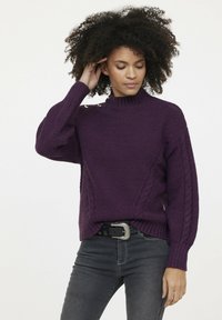 Pull en laine violet avec un motif texturé, un col côtelé et des accents en tricot torsadé sur les manches. Assorti à un jean foncé et une ceinture.
