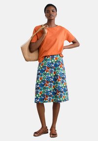 Orange linen t-shirt paired with a floral-patterned skirt in blue, green, orange, and white. Model holds a woven bag and wears brown sandals.