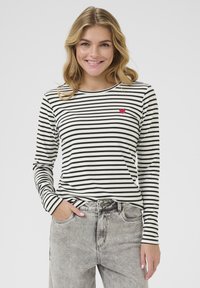 Camicia a maniche lunghe a righe in crema e nero con un dettaglio a forma di cuore rosso sul petto, abbinata a jeans grigi a vita alta.