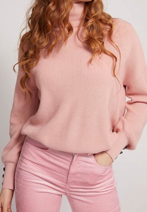 Frau mit langen, lockigen roten Haaren, die einen rosa gerippten Rollkragenpullover und rosa Cordhosen trägt, mit einer Hand in der Tasche.