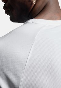 Biała sportowa koszulka z teksturowanej tkaniny w kratkę. Posiada okrągły dekolt i raglanowe rękawy, odsłaniając część szyi i ramienia.