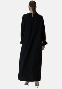 Langes schwarzes Kleid mit einem Schlüsselloch-Rücken, langen Ärmeln und gerafften Manschetten. Glattes, fließendes Material und eine lockere Silhouette.