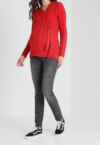 Punainen palmikkoneuleinen äitiysvillapaita, jossa on V-kaula-aukko ja vyötärölle sidottava nauha. Yhdistetty harmaisiin kapeisiin farkkuihin ja mustiin tennareihin. Sujuva pinta, rento istuvuus.