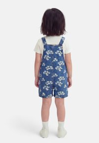 Enfant aux cheveux foncés, vu de dos, portant une chemise blanche, une salopette courte en jean bleu à fleurs, des chaussettes blanches et des baskets blanches sur fond blanc.