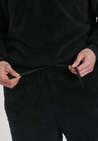 Schwarzes, flauschiges Fleece-Loungewear-Set mit sichtbarer Schleifenstruktur; das Modell hält den schwarzen Kordelzug an der Taille. Lange Ärmel, lockere Passform.