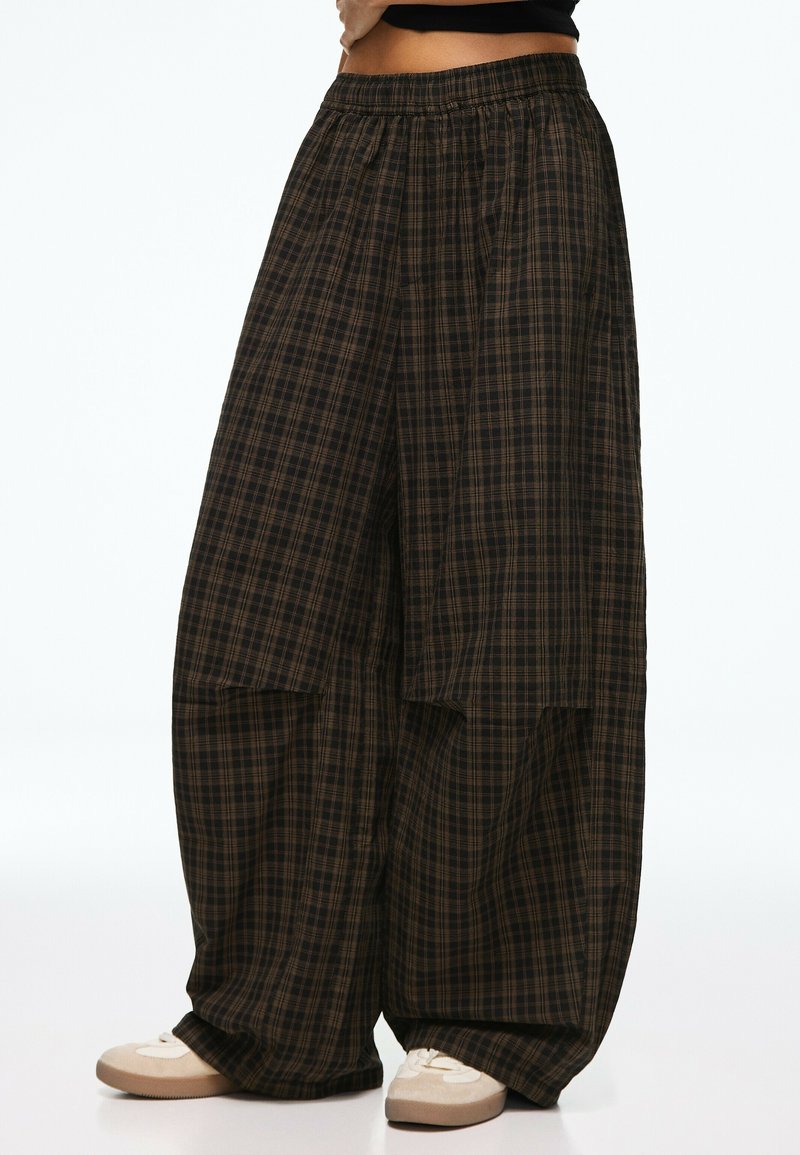 Pantalon large à carreaux marron et noir avec taille élastique, porté avec des baskets blanches et un haut noir.