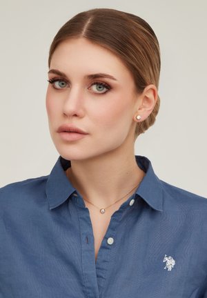 Femme à la peau claire et aux yeux verts, portant une chemise bleue à col, des boucles d'oreilles en diamant en forme de clou et un collier assorti avec un pendentif en diamant.