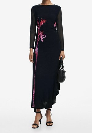 Woman wearing a black long-sleeve dress with pink floral design, holding a black handbag, and wearing black high-heeled sandals.