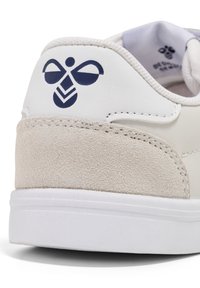 Vit lädersneaker med en beige mocka-häl, med en marinblå logotyp. Slät ovandel med kontrasterande texturer och en vit sula.