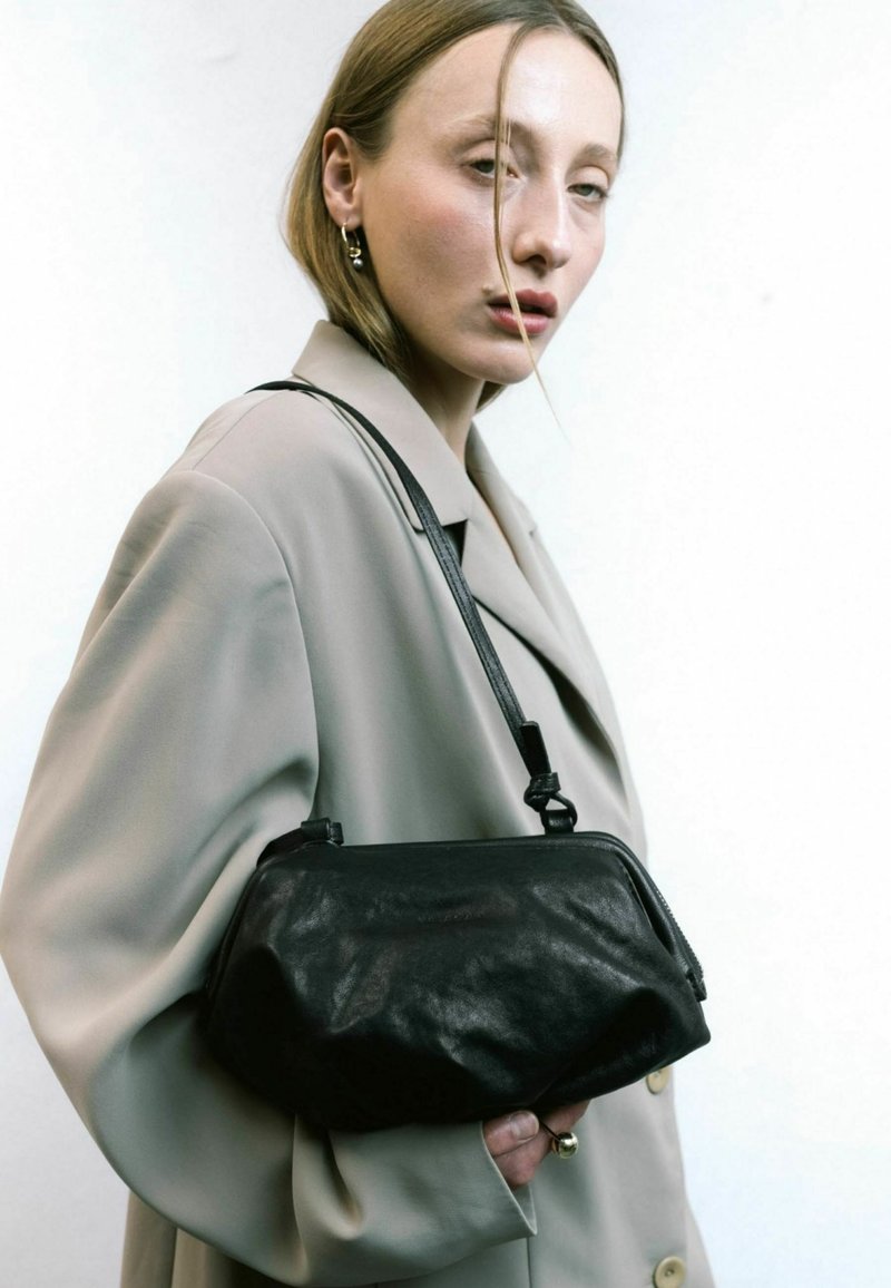 Bolso de mano de cuero negro con forma curva, una correa corta y una superficie texturizada, sostenido contra un conjunto y fondo neutros claros.