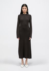 Długa, dopasowana brązowa sukienka z długimi rękawami i wysokim dekoltem. Materiał jest gładki z lekkim streczem, posiada boczne rozcięcia dla swobody ruchu.