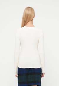 Top cu mânecă lungă, cu model în dungi, de culoare crem, având un decolteu ondulat și o siluetă mulată, asortat cu o fustă în carouri albastre și verzi.