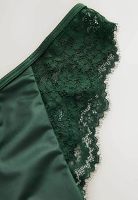 Lenjerie intunecată verde, realizată dintr-un material satinat neted, cu detalii delicate din dantelă. Dantela are motive florale și o margine zimțată.